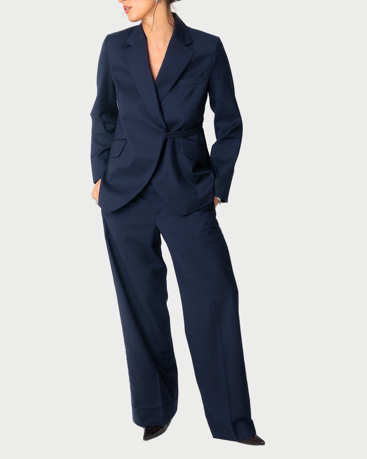 A woman models the WE-AR4 Smoking Blazer, a dark navy upcycled virgin wool pantsuit by WE-AR4 with wide-leg trousers, standing against a plain light background, hands in her pockets and head turned slightly to the side.