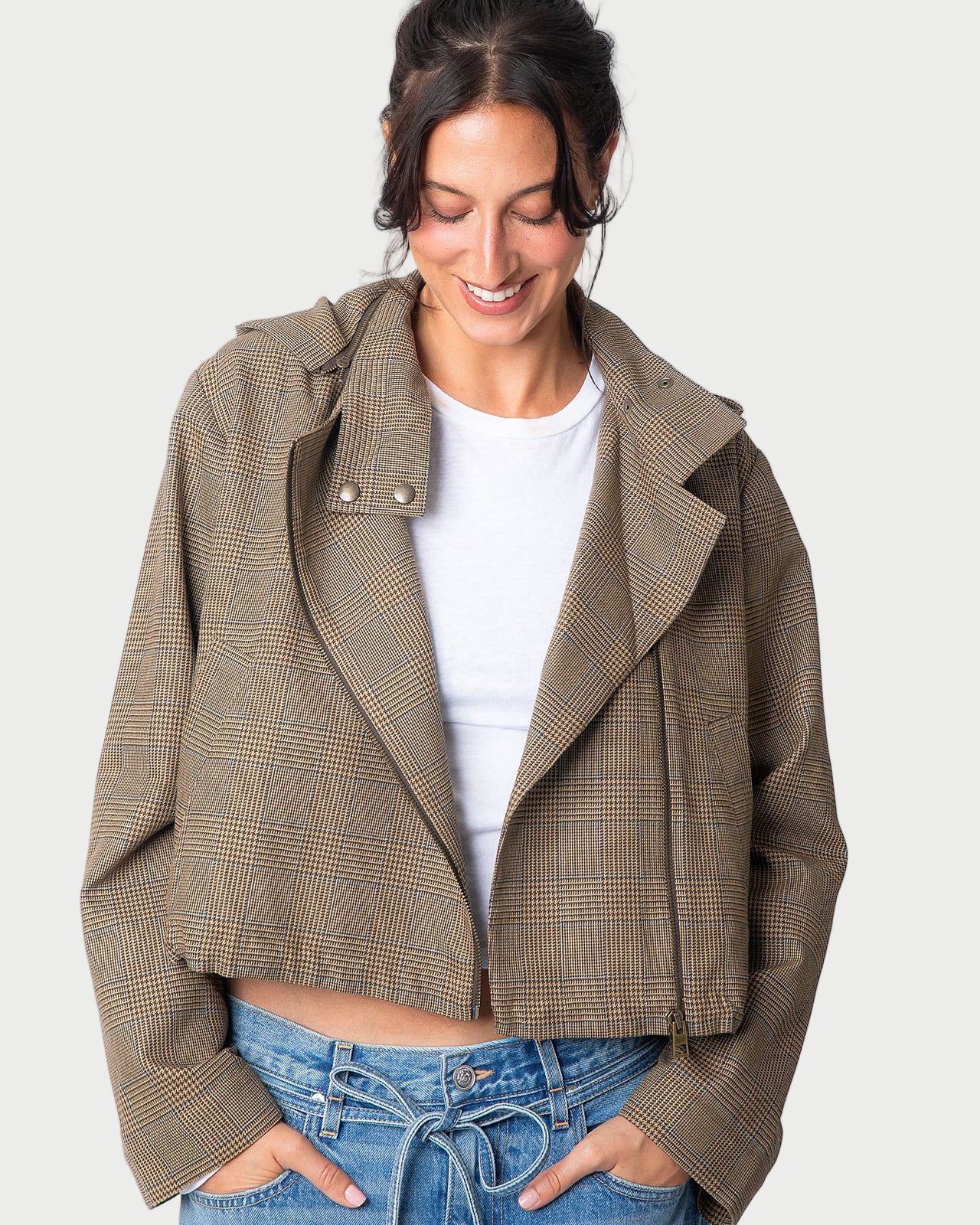 A woman smiles as she looks down, hands in her pockets, wearing the WE-AR4 Underground Bomber Jacket by WE-AR4 in light brown plaid over a white t-shirt and blue jeans.