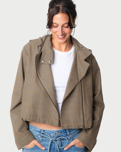 A woman smiles as she looks down, hands in her pockets, wearing the WE-AR4 Underground Bomber Jacket by WE-AR4 in light brown plaid over a white t-shirt and blue jeans.