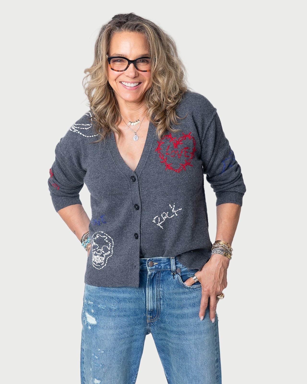 A smiling woman with wavy light brown hair and glasses wears the Zadig & Voltaire Mirkaz Cardigan, standing with her hands in her pockets against a white background.