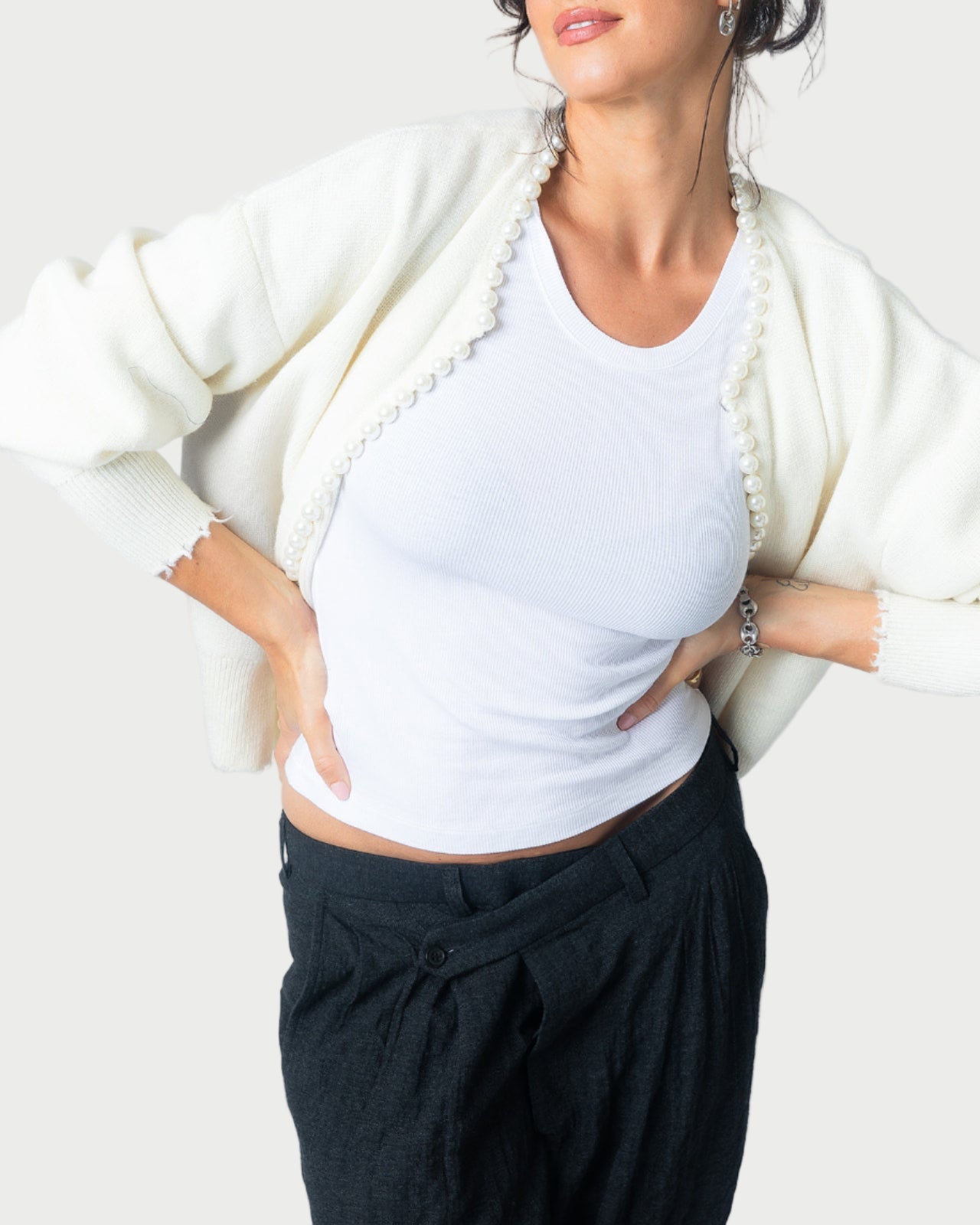 A woman poses confidently with her hands on her hips against a light background, wearing the Essentiel Antwerp Ideas Beaded Cardigan over a white tank top and black pants.