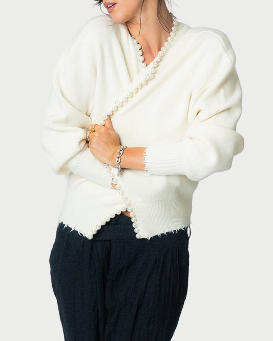 A woman wears Essentiel Antwerp's Ideas Beaded Cardigan with black pants, holding a pearl necklace. Her head is slightly tilted and her hair is loosely tied back, highlighting the cozy charm of this knitwear piece.