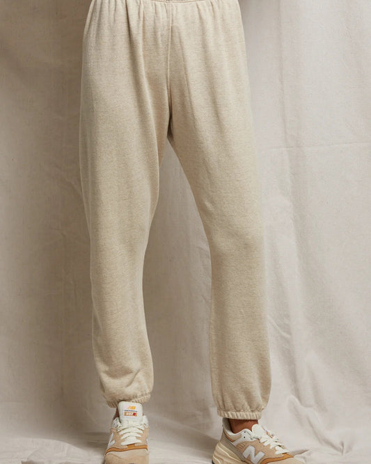 A person wears perfectwhitetee Johnny Fleece Sweatpant in heather grey, paired with tan New Balance sneakers, standing against a light fabric backdrop. The image shows only their lower body, cropped at the waist.