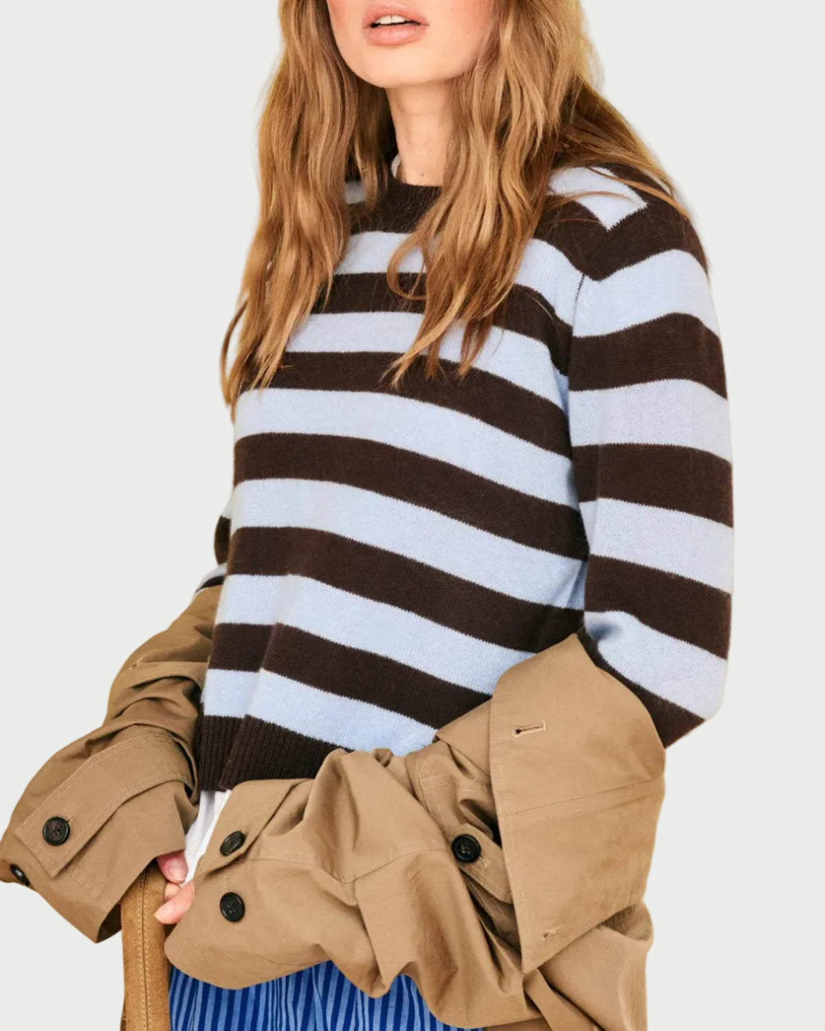 A person with long wavy hair stands against a plain, light-colored background wearing the JUMPER1234 Cashmere Stripe Crew, a lightweight cashmere sweater in light blue and dark brown stripes with a crew neck. They pair it with a pleated blue skirt and hold a tan coat.