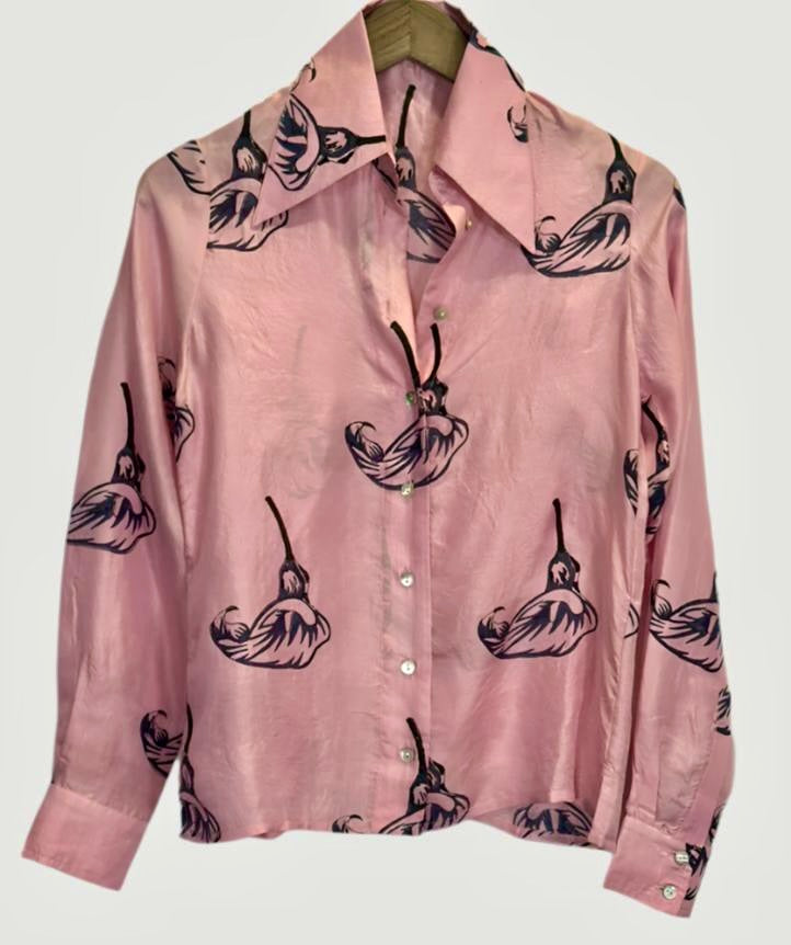 The Pink Calla Lily Shirt by Alix of Bohemia is a pink silk blouse adorned with a delicate pattern of pink calla lilies and black abstract swans. It features a large collar and is elegantly buttoned in the front with white buttons, gracefully displayed on a wooden hanger against a minimalist backdrop.