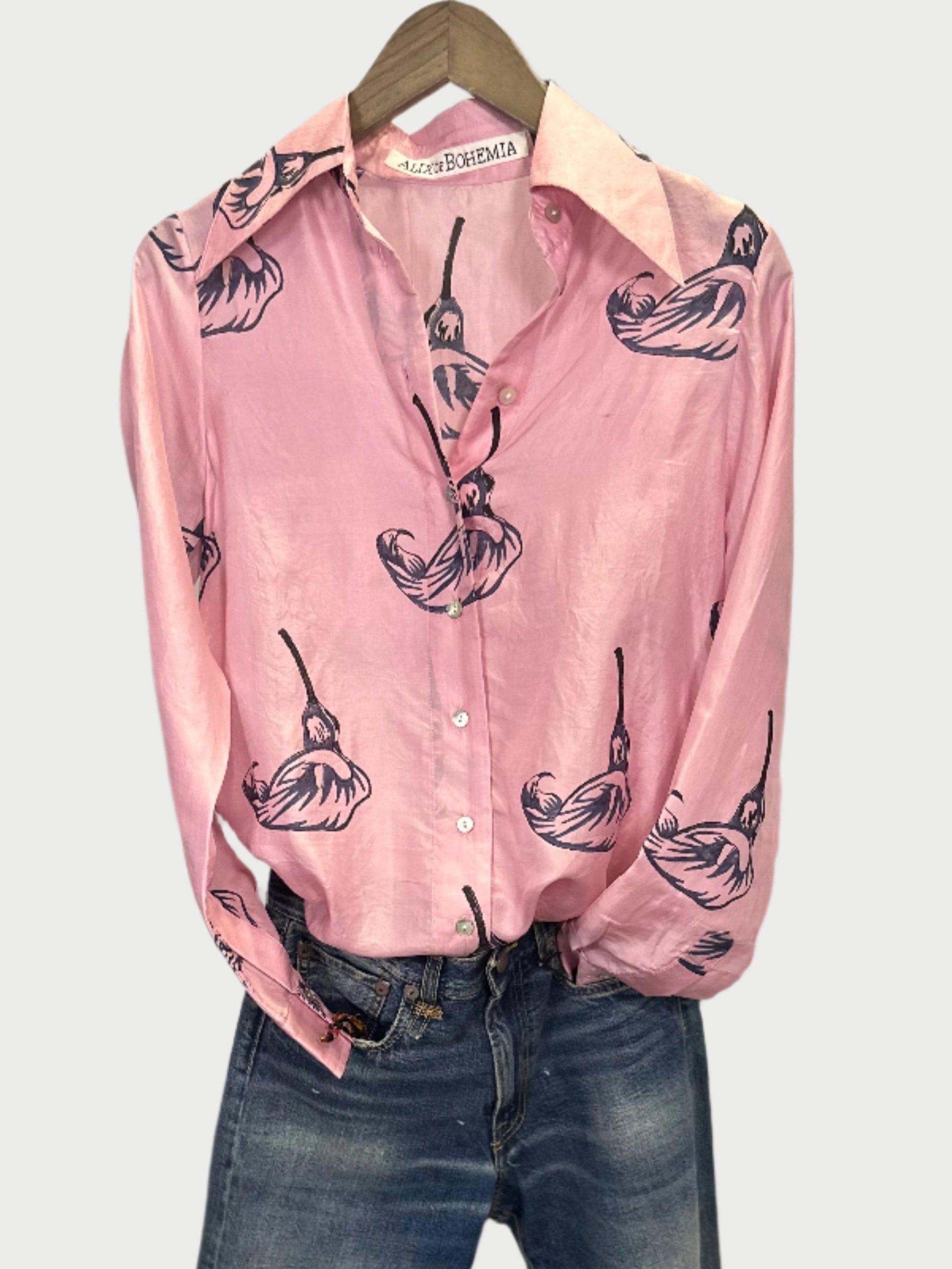 A Pink Calla Lily Shirt from Alix of Bohemia is showcased on a hanger over denim blue jeans. This silk blouse features a charming calla lily pattern, complete with a collar and front buttons, enhancing its elegant floral design.
