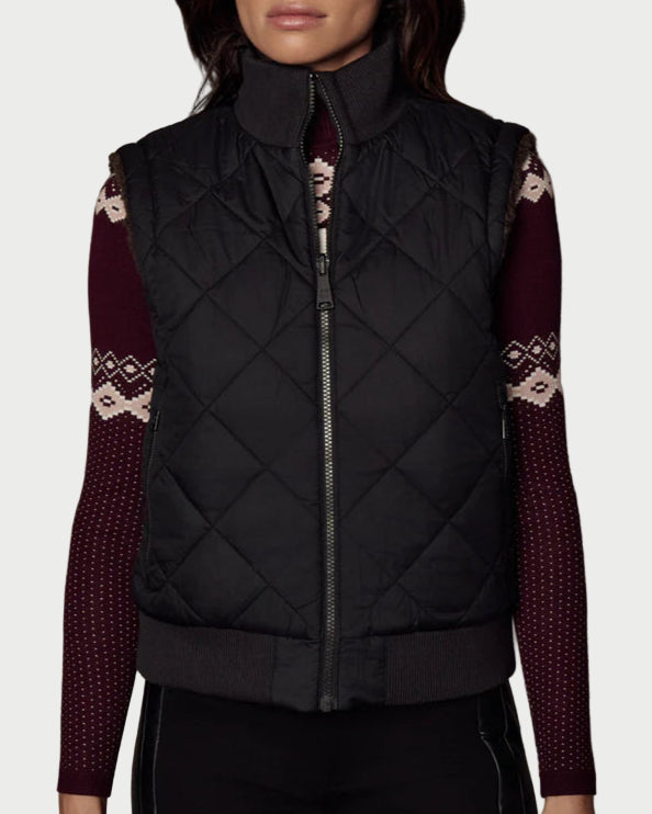 Woman wearing a black quilted vest over a patterned long-sleeve shirt against a textured gray background