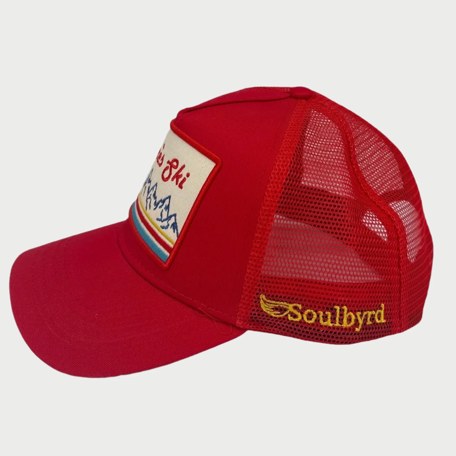 The Soulbyrd Apres Ski Red Trucker Hat features a curved bill, red mesh, mountain patch with “Ski” on the front, and “Soulbyrd” embroidered in yellow on the side—a must-have from Soulbyrd for any ski lodge trip.