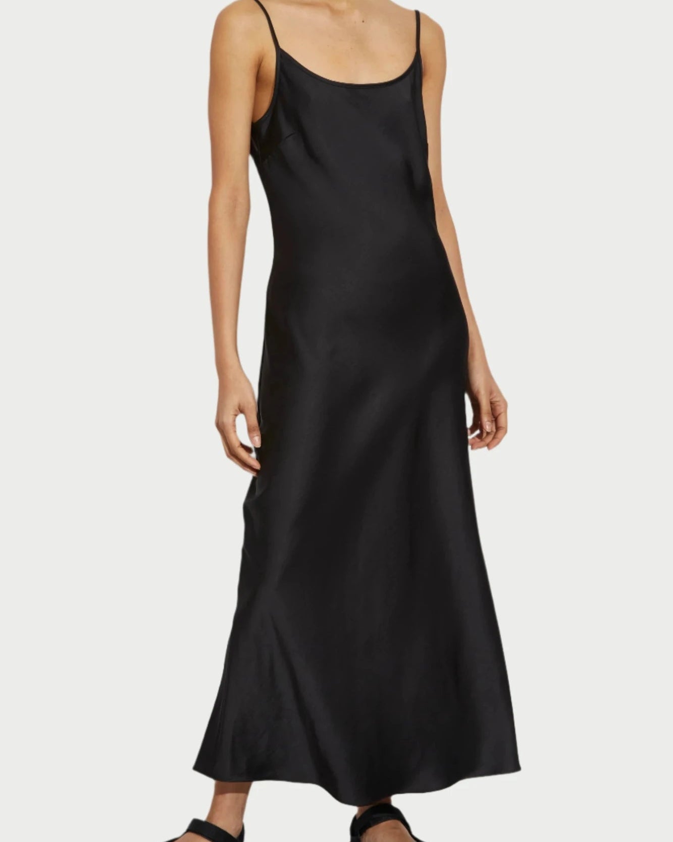 A person stands against a white background, wearing the Black Haley Slip Dress by Saint Art NY—a long, simple black slip dress with thin straps. They have dark hair pulled back and wear black sandals. The person's expression is neutral, and their hands rest by their sides.