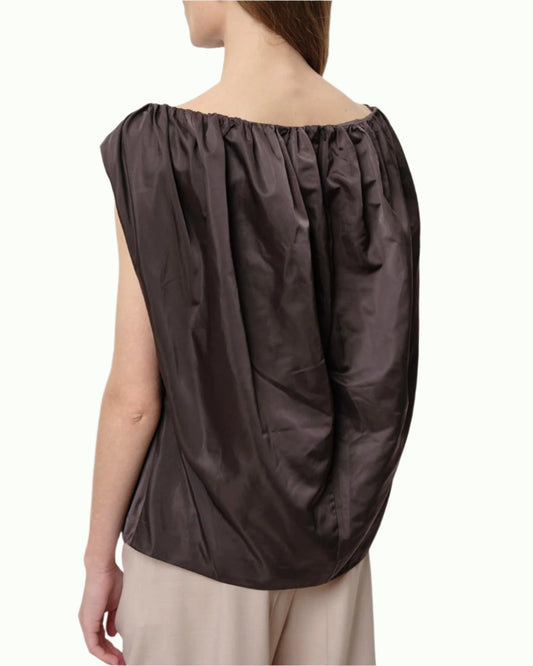 A woman wears the Herskind Martina Top, a loose dark brown sleeveless top with a gathered neckline and draped back, paired with light beige pants for effortless cocktail style against a plain light background.