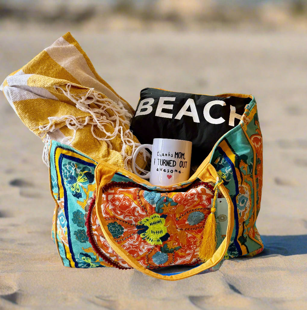 The DEBBIE KATZ Boho Bag, shown indoors near a window and clothes, holds a yellow-and-white towel, a "BEACH" shirt, and a mug that says "Thanks Mom, I turned out awesome.