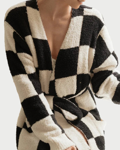 Wearing the LOCAL BEACH Black Checker Luxe Robe, a person sits comfortably with one arm bent and the other on their leg. This plush microfiber robe from LOCAL BEACH features a bold checkerboard pattern and is belted at the waist for ultimate comfort.