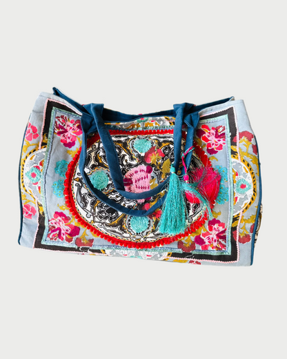 The DEBBIE KATZ Boho Bag is an artisanal masterpiece with colorful embroidery featuring vibrant floral and geometric patterns. It has blue handles, plus teal and pink tassels, making it perfect for a beach day.