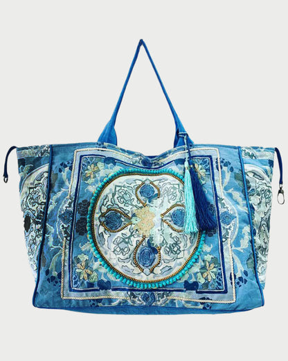 The large blue Boho Bag by DEBBIE KATZ is ideal for the beach, featuring intricate artisanal embroidery with circular and floral motifs in blue, green, and white. It comes with thin shoulder straps and a front decorative tassel.