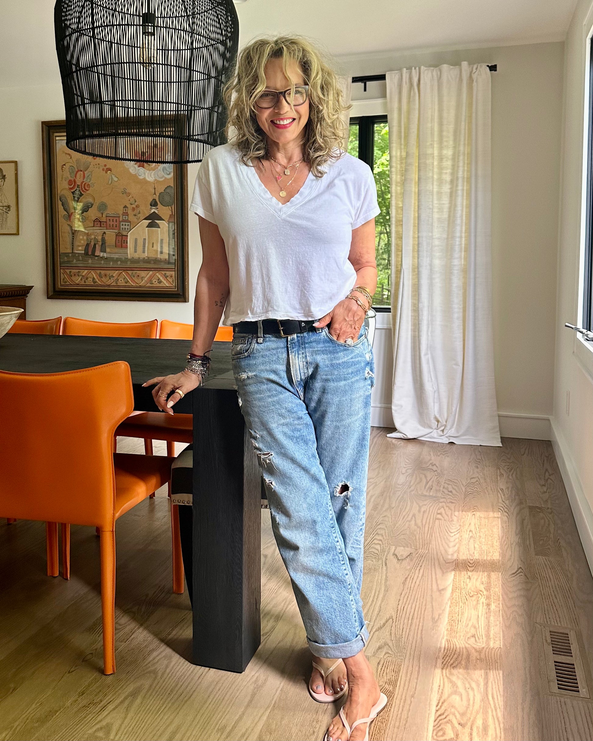 A woman with curly blonde hair and glasses smiles in a bright dining room, wearing a white T-shirt, R13 Boyfriend Jean In Bain Blue by R13, and flip-flops as she leans against a dark table with orange chairs.
