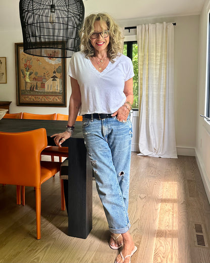 A woman with curly blonde hair and glasses smiles in a bright dining room, wearing a white T-shirt, R13 Boyfriend Jean In Bain Blue by R13, and flip-flops as she leans against a dark table with orange chairs.