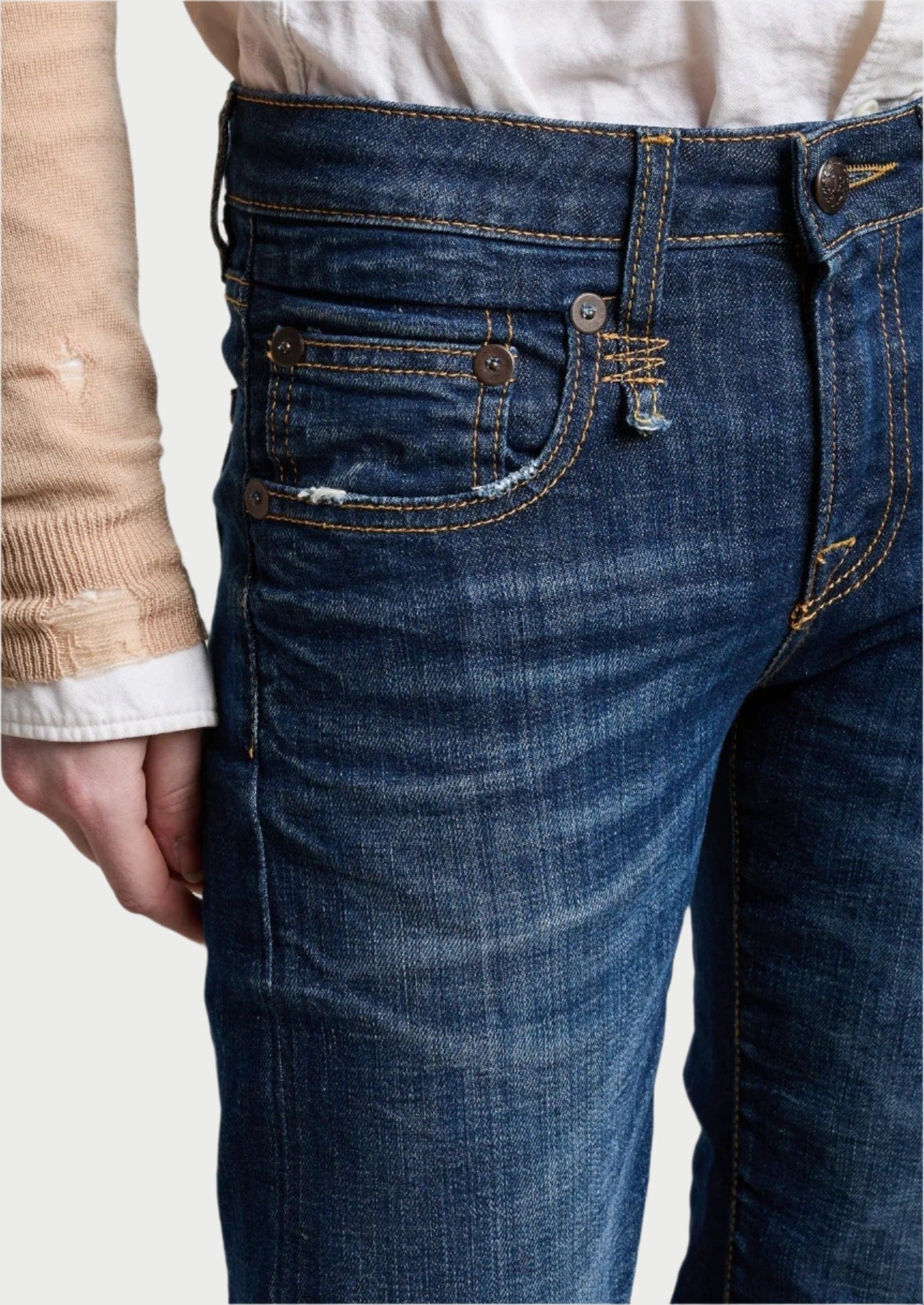Close-up photo of a person wearing blue R13 Boy Straight Jeans with a frayed white sleeve peeking out under a tan sleeve. The stretch and rigid denim features multiple pockets, contrasting stitching, and visible wear marks. The background shows a textured, white-painted brick wall.