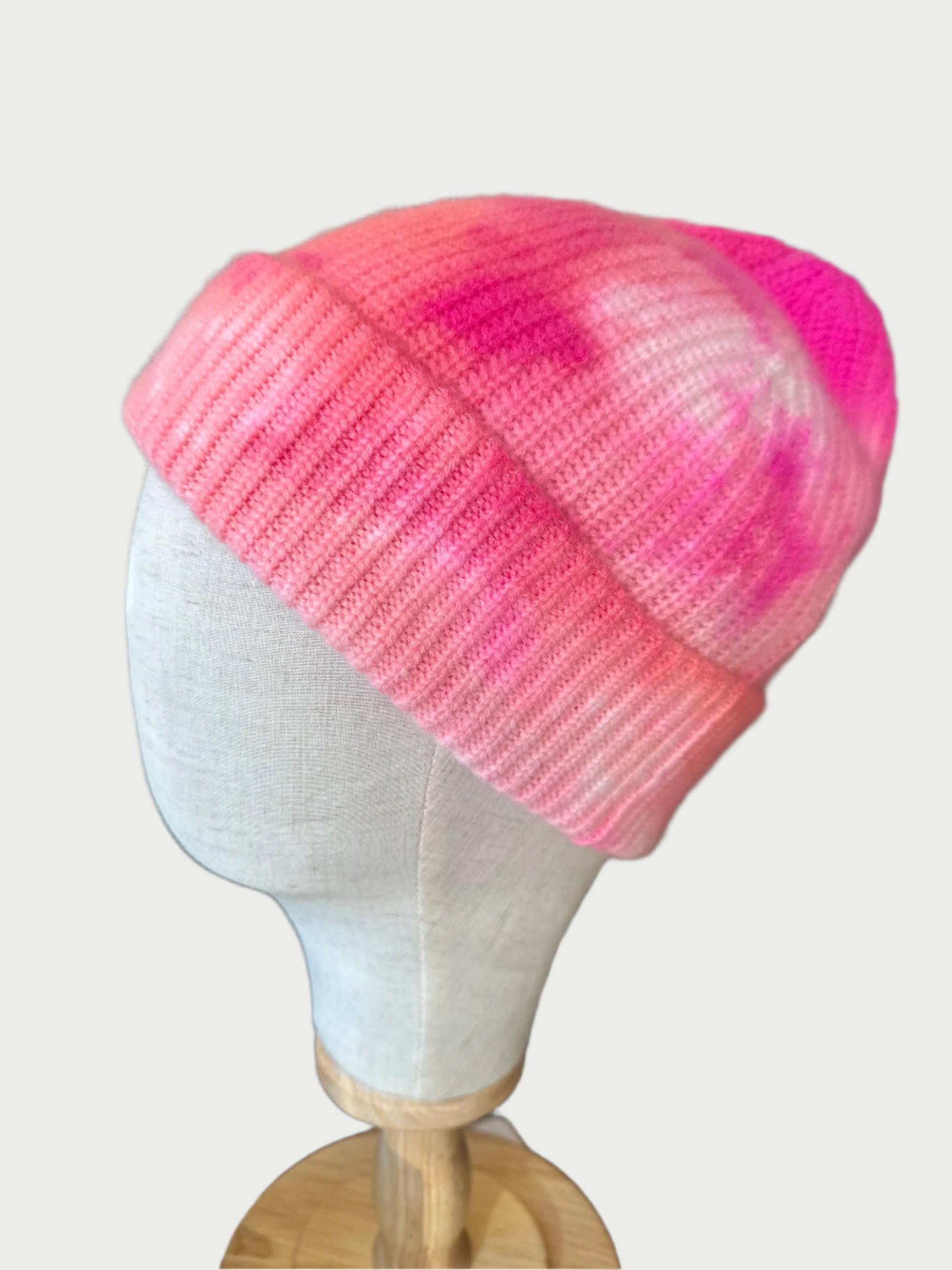 A mannequin head displays the pink Dip Dyed Beanie by MAREA, with an artisanal dye creating charming pink and white tie-dye patterns. The backdrop is a store interior with hat-lined shelves, highlighting a curated collection of styles.