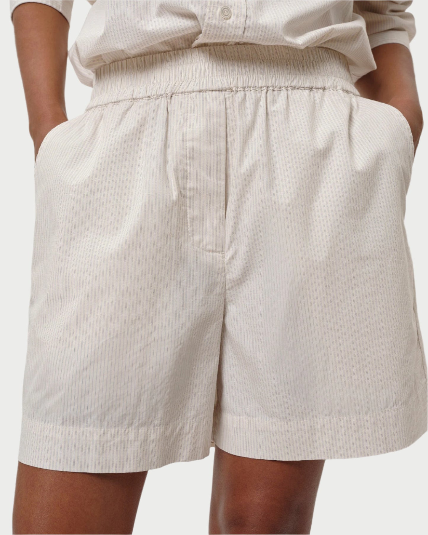 An individual is sporting the Alma Shorts by Herskind. These light-colored, 100% cotton shorts feature vertical stripes, a relaxed-fit with an elastic waistband, and pockets for a casual look. They pair perfectly with a matching button-up shirt as their hands rest in the pockets.