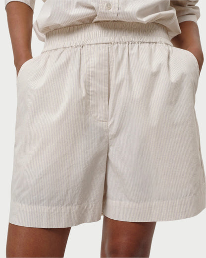 An individual is sporting the Alma Shorts by Herskind. These light-colored, 100% cotton shorts feature vertical stripes, a relaxed-fit with an elastic waistband, and pockets for a casual look. They pair perfectly with a matching button-up shirt as their hands rest in the pockets.