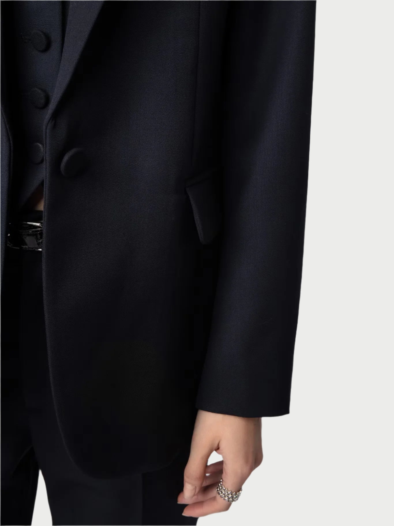 Dressed in a tailored navy blazer by Zadig & Voltaire, a person wearing a sleek black suit and coat stands with their ring-adorned hand resting at their side against a plain light background.