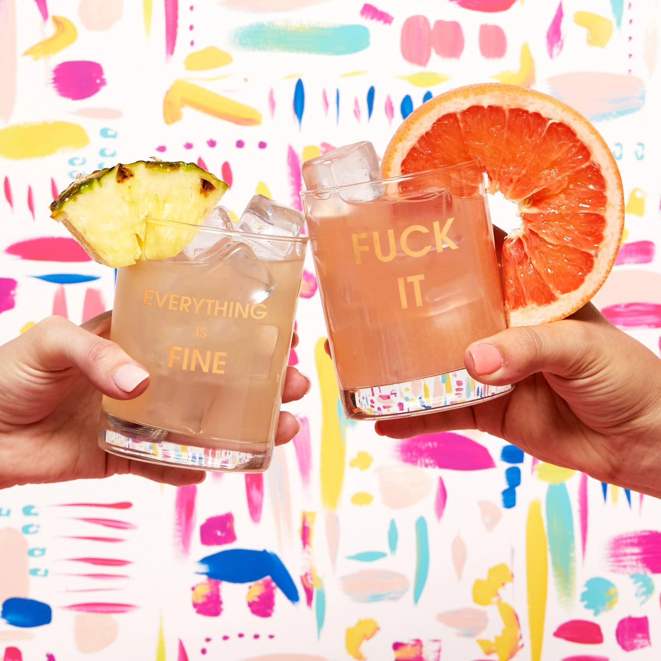Two hands holding cocktails against a colorful abstract background. The left glass, enhanced with a pineapple garnish, says "EVERYTHING is FINE" in gold foil. The right rocks glass by Chez Gagné features a grapefruit slice and reads "FUCK IT.