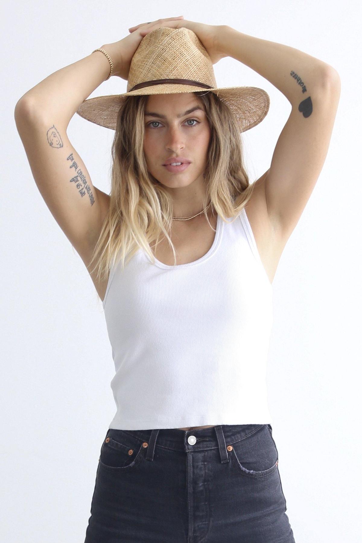 A person in a straw hat, the perfectwhitetee Blondie Tank by perfectwhitetee, and black pants poses with arms raised, displaying tattoos on both arms against a plain white background.
