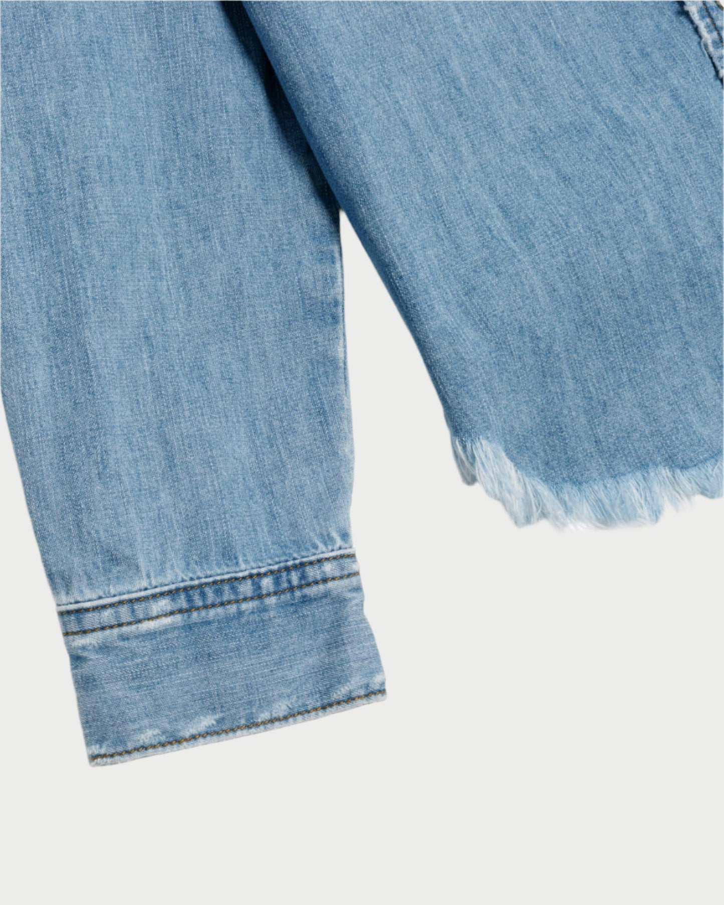 Close-up of the RE/DONE Western Denim Shirt, a rodeo staple with frayed hem and cuff on a pale background. The texture and stitching highlight the casual, worn look of cotton chambray.