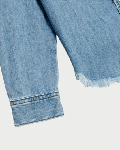 Close-up of the RE/DONE Western Denim Shirt, a rodeo staple with frayed hem and cuff on a pale background. The texture and stitching highlight the casual, worn look of cotton chambray.