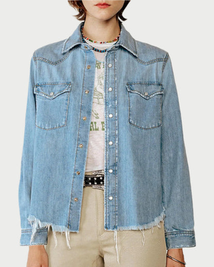 The person dons a RE/DONE Western Denim Shirt with snap buttons and frayed edges over a white graphic T-shirt. Two chest pockets enhance the light blue denim, paired with a beaded necklace and beige pants for an effortless rodeo staple vibe.