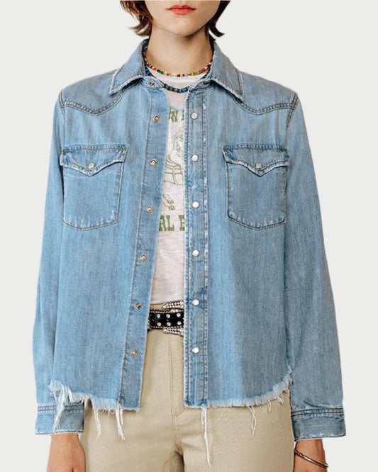 The person dons a RE/DONE Western Denim Shirt with snap buttons and frayed edges over a white graphic T-shirt. Two chest pockets enhance the light blue denim, paired with a beaded necklace and beige pants for an effortless rodeo staple vibe.