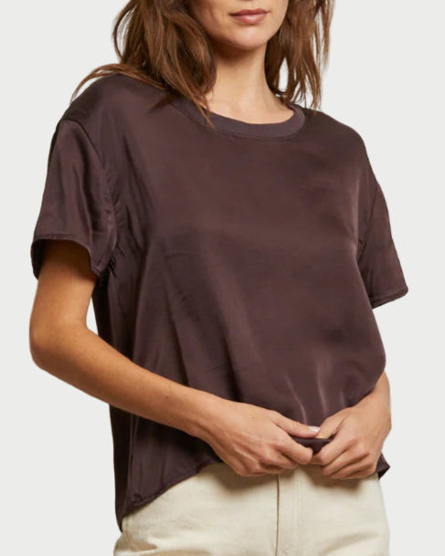 A woman wearing the Delphine Satin Crewneck Tee in Cocoa by perfectwhitetee and light-colored pants stands against a plain white background.