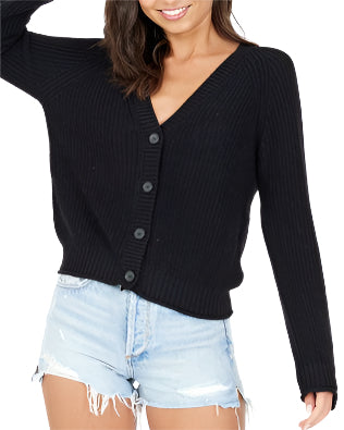 A person with dark hair is smiling and looking down while posing in a black oversized Raleigh Cardigan by One Grey Day, made of cotton fabric, paired with frayed light denim shorts against a plain white background.