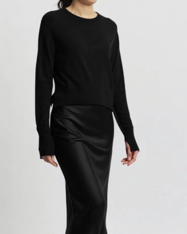 A woman wears the Saint Art NY Elodie Sweater Black, a long-sleeve merino wool top, and a long black satin skirt, standing against a plain light gray background. Her head is partially out of frame.