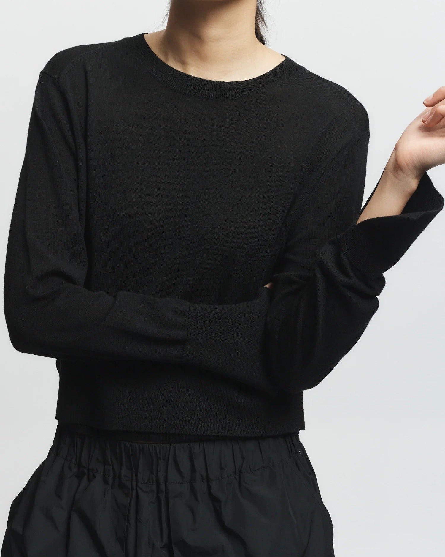 Wearing the Saint Art NY Elodie Sweater in black and a black skirt, a person stands against a plain light background, one arm bent and the other resting across their body, with their face partially out of frame.