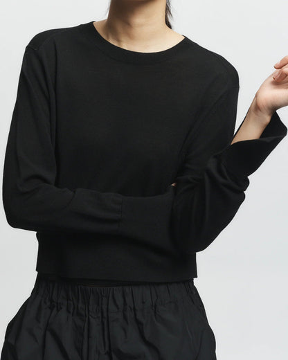 Wearing the Saint Art NY Elodie Sweater in black and a black skirt, a person stands against a plain light background, one arm bent and the other resting across their body, with their face partially out of frame.