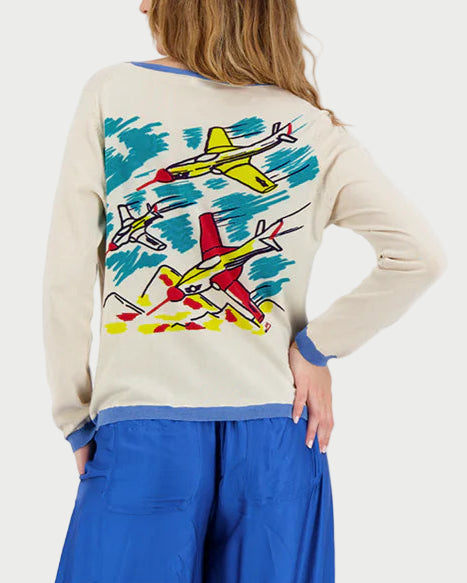 Woman wearing a cream sweater with colorful airplane designs and blue wide-leg pants on a white background