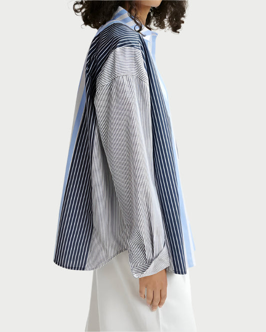 With curly hair, a person dons an oversized, lightweight Essentiel Antwerp Patchwork Shirt featuring vertical blue and white stripes, effortlessly paired with white pants. The cotton blend fabric drapes elegantly as they stand against a light background, slightly turned to the side.