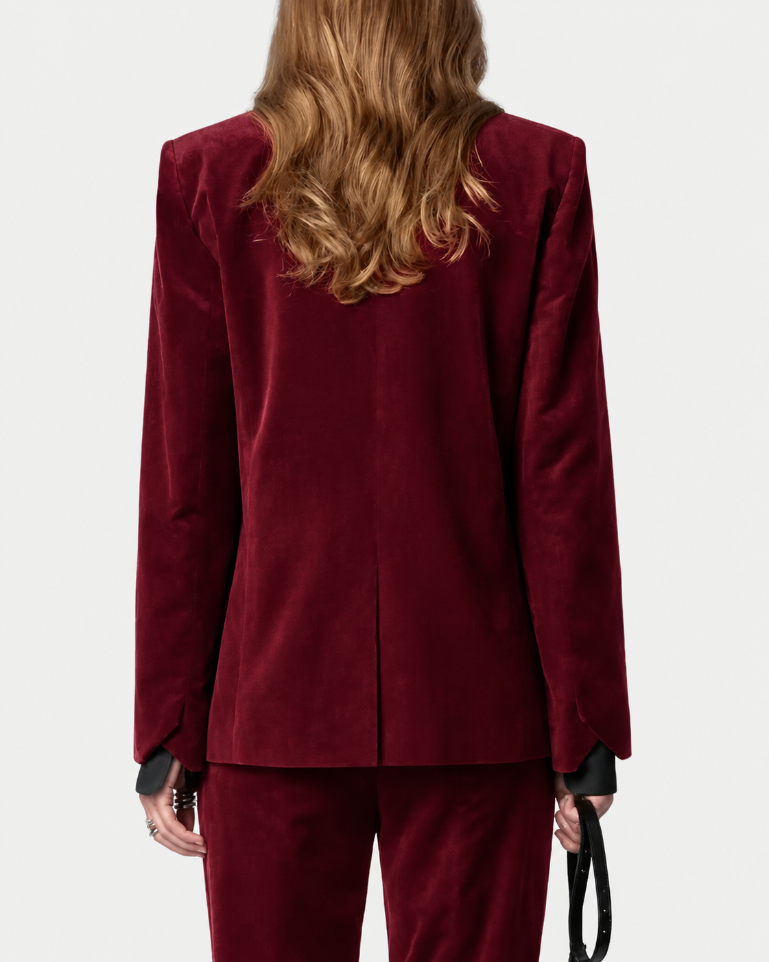 A person with long, wavy light brown hair is shown from the back wearing the Zadig & Voltaire Vavy Velvet Blazer and matching pants set in burgundy cotton velvet. Their left hand holds a thin black strap, and the background is plain and light-colored.