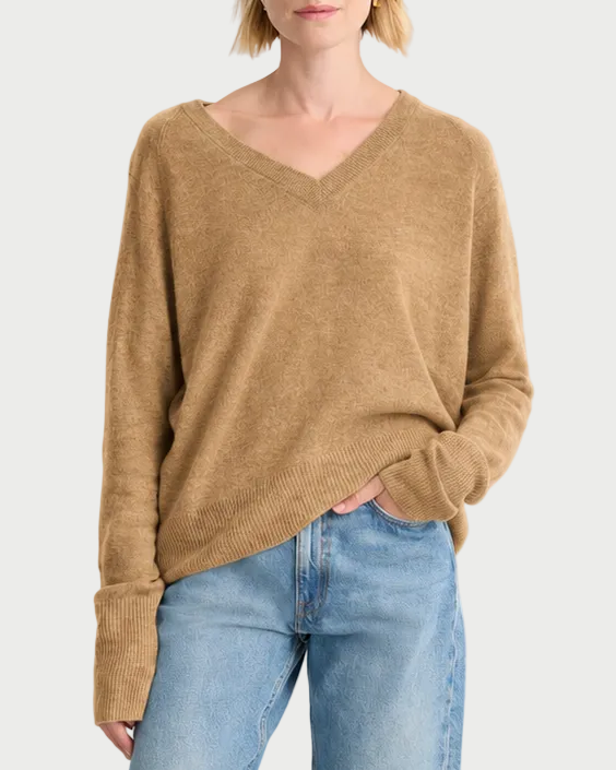 A woman modeling a light brown v-neck knit sweater and light-wash blue jeans against a plain white studio background.