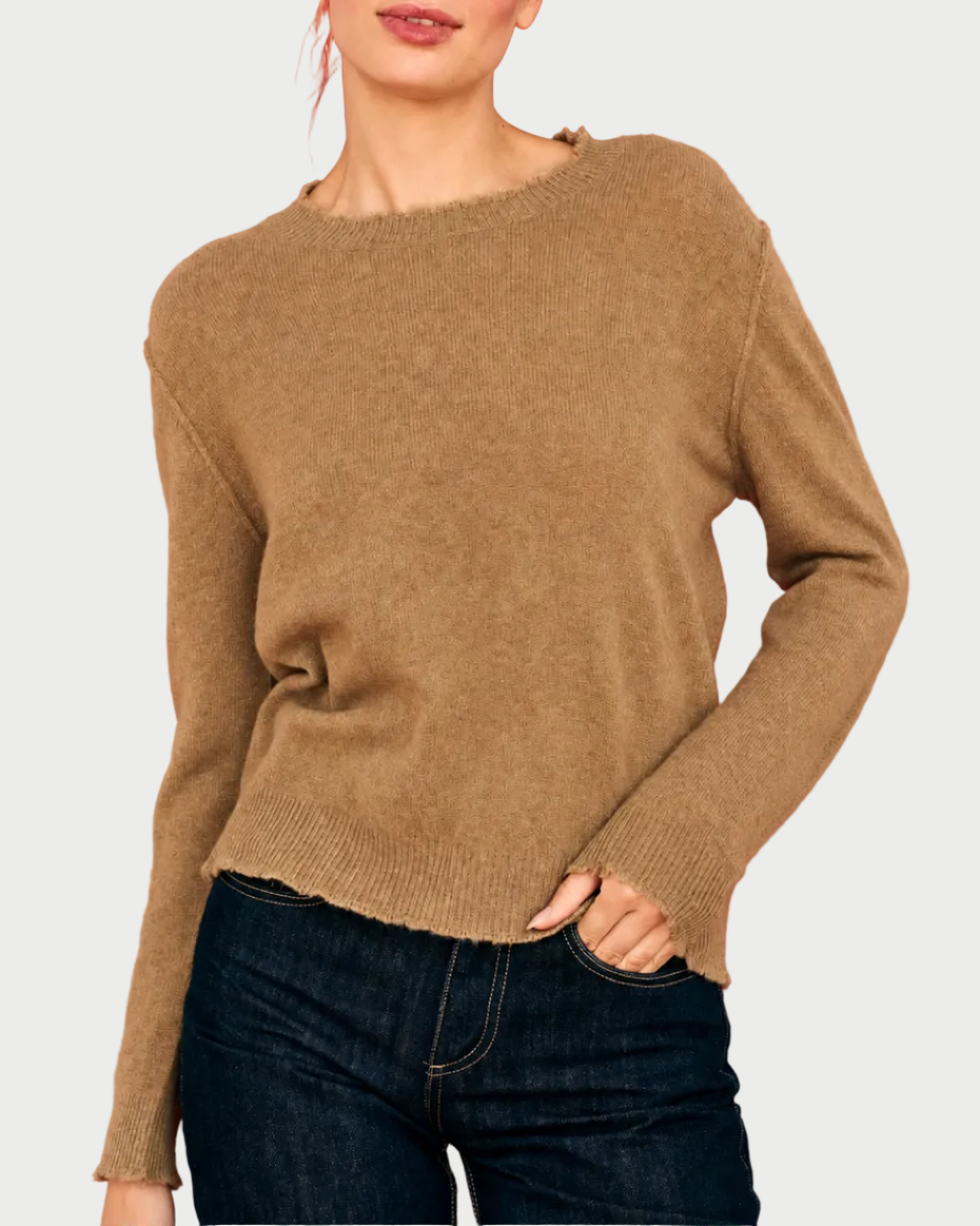 A woman wears the JUMPER1234 Distressed Cashmere Crew Sandstone, a relaxed fit knit sweater with distressed edges and long sleeves, styled with dark blue jeans against a plain background.