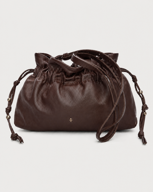 The Henry Beguelin Floriana Clutch by Henry Beguelin is a dark brown luxury handbag crafted from supple Italian leather, featuring a gathered top, long adjustable straps, and elegant gold bead accents. A small gold emblem decorates the bottom center of this handcrafted bag, displayed against a plain light background.