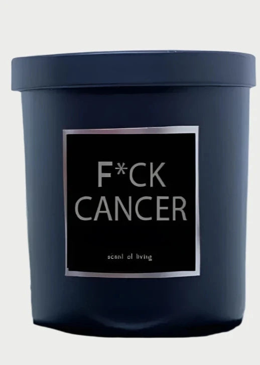 A black candle jar from Rude Dude with a label that reads "F*ck Cancer" in bold white text, and "scent of living" in smaller text below it. Made in the USA, this 9oz jar is set against a blurred background featuring an extended middle finger sculpture or hand gesture.