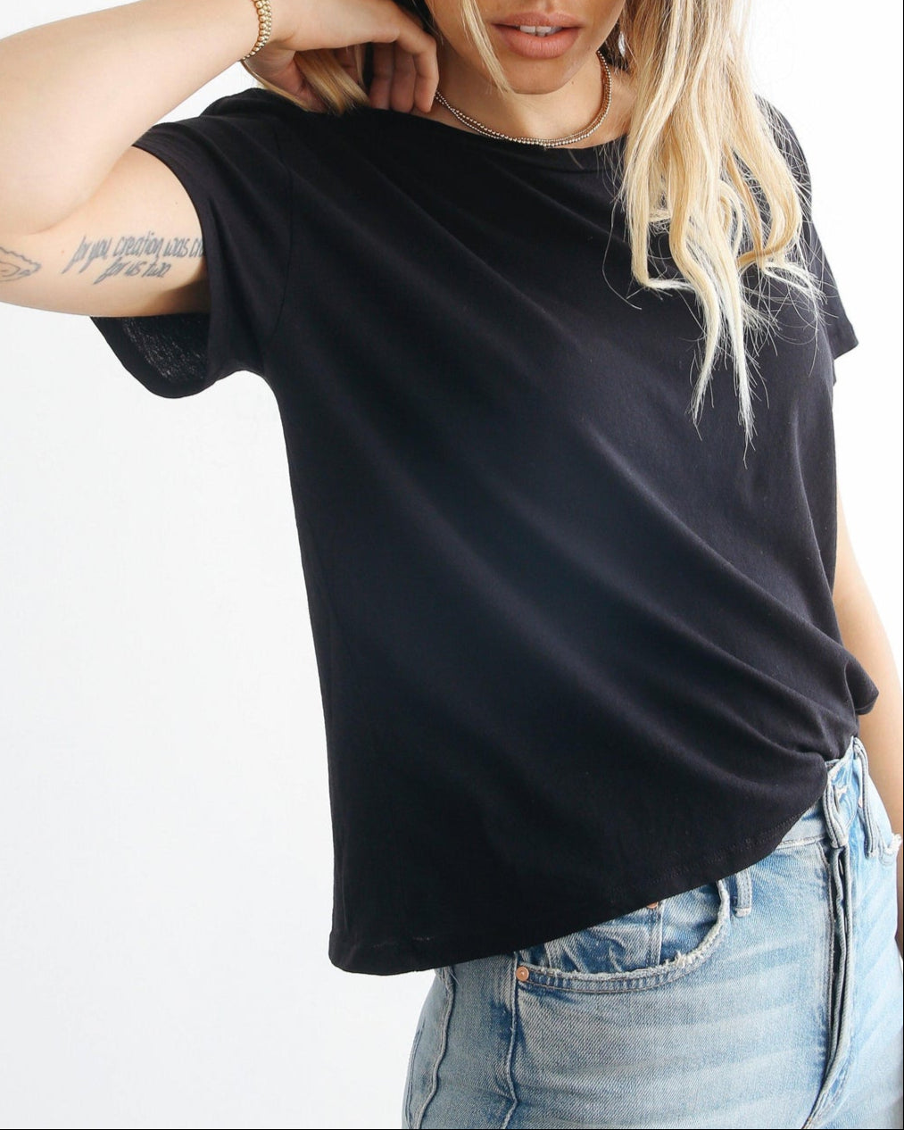 A woman with wavy blonde hair poses against a white background wearing light blue jeans and the Harley Boxy Crew in Black and White by perfectwhitetee. She rests one hand on her neck, revealing a tattoo on her forearm, while showcasing the tee's versatility as an ideal layering piece.