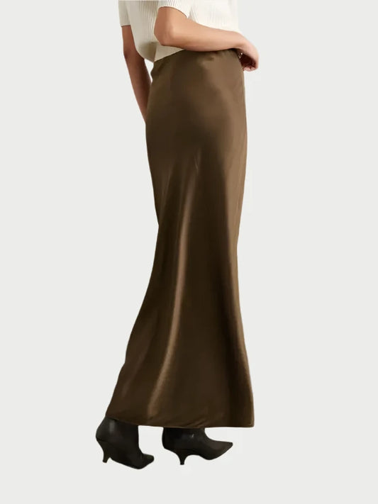 A person is dressed in the Silk Column Maxi Skirt by Frame, teamed with black ankle boots and a white, short-sleeved top. They are posed against a solid light gray background, exhibiting a column silhouette while facing sideways to the left.