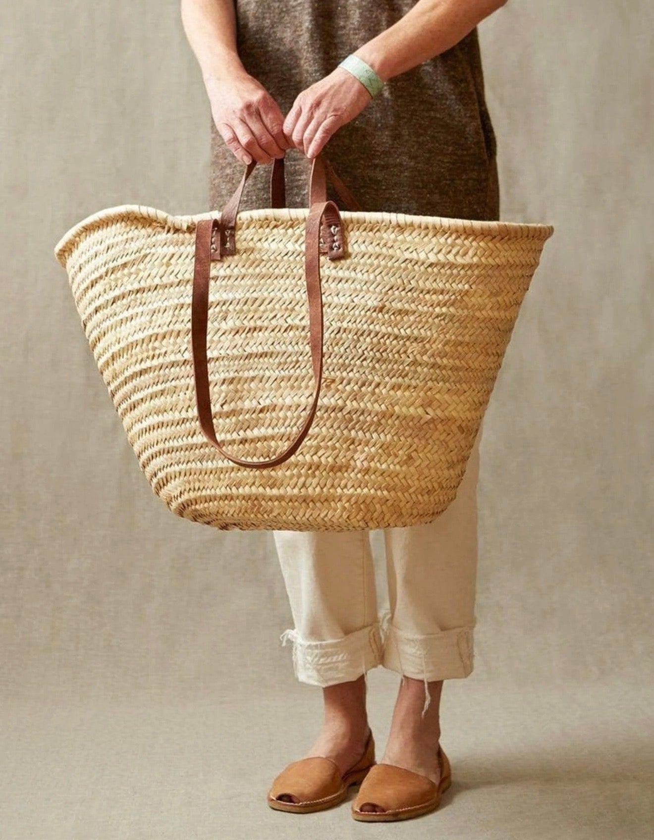 A person holds the WEST X WEST Straw Tote by West X West, featuring brown leather handles, while wearing a brown top, off-white pants, and brown sandals against a neutral background.