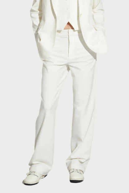 A person stands against a plain background wearing a white suit from Saint Art NY. The ensemble, featuring the Cait Low-Rise Tuxedo Pants along with a blazer and waistcoat in breathable fabric, is paired with white shoes. With hands in pockets and a neutral expression, it's the epitome of modern tuxedo dressing.