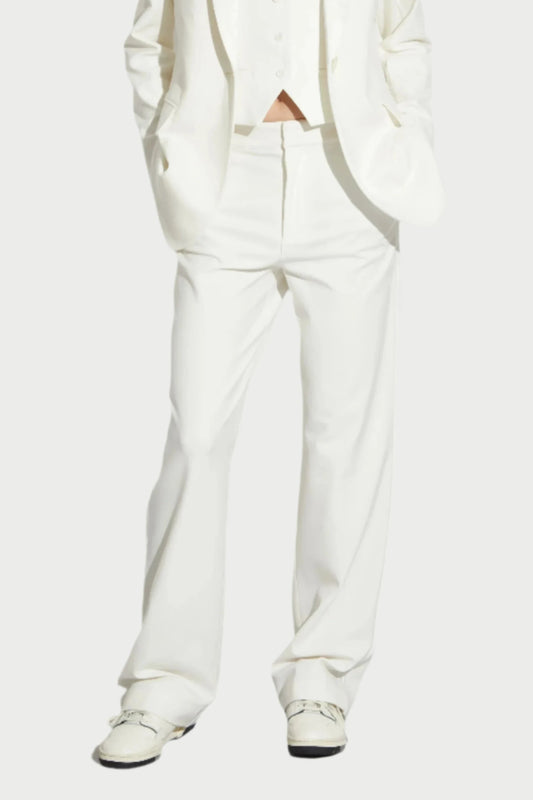 A person stands against a plain background wearing a white suit from Saint Art NY. The ensemble, featuring the Cait Low-Rise Tuxedo Pants along with a blazer and waistcoat in breathable fabric, is paired with white shoes. With hands in pockets and a neutral expression, it's the epitome of modern tuxedo dressing.