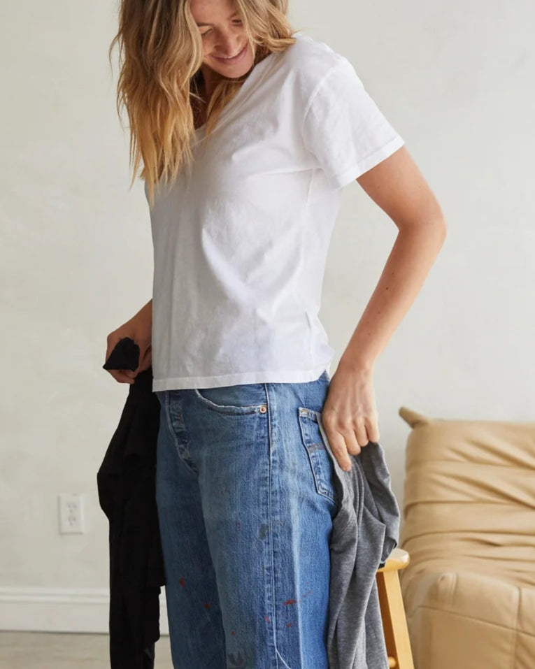 A person with long hair, wearing a Hendrix Basic V-Neck T-Shirt by perfectwhitetee and blue jeans, holds a black and gray garment. Next to them is a light-colored chair in a softly lit room, embodying the essence of a wardrobe staple.