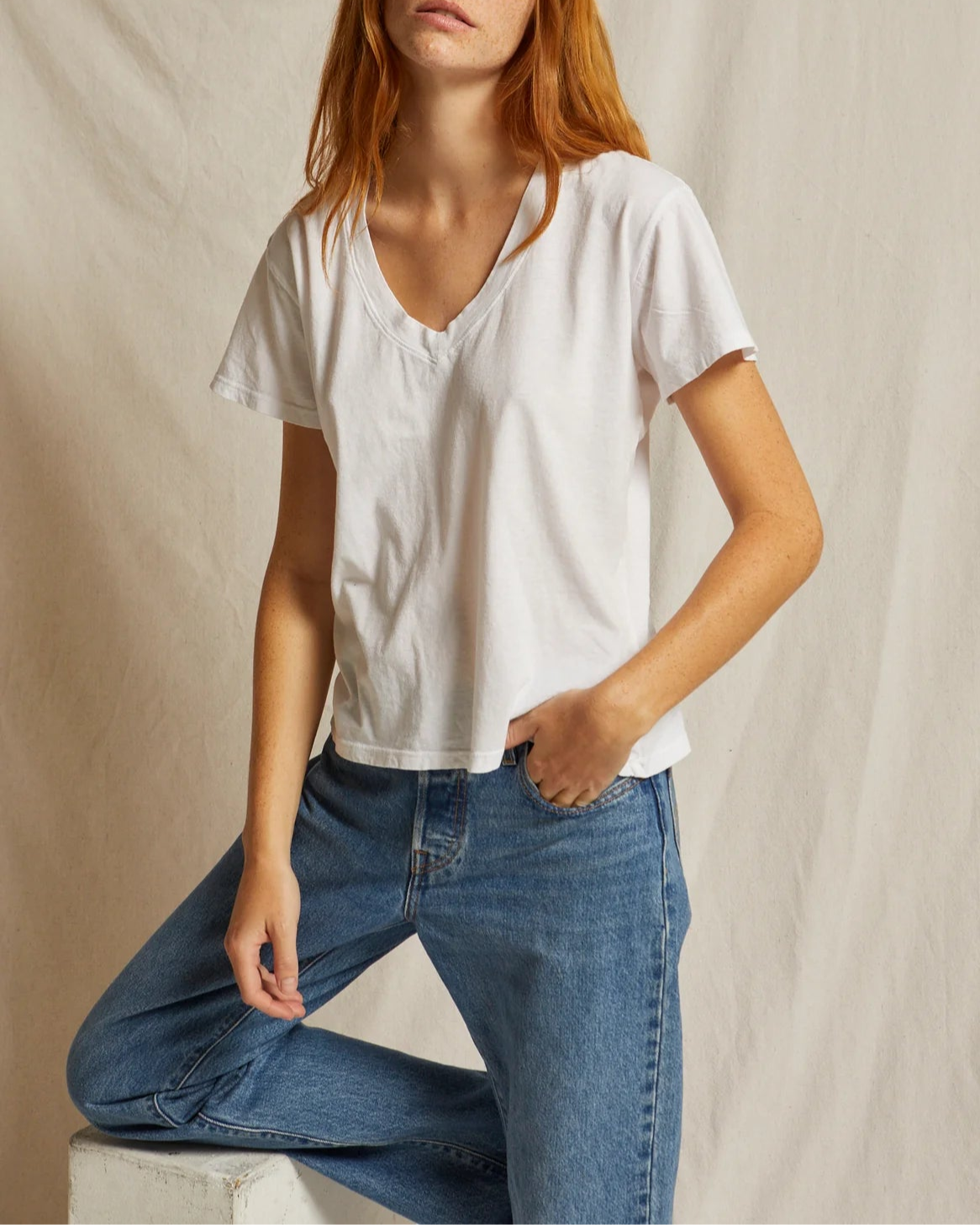A person with long red hair is wearing a Hendrix Basic V-Neck T-Shirt White by perfectwhitetee and blue jeans, standing against a plain light-colored backdrop. They lean forward slightly with one hand in their pocket, highlighting this essential wardrobe piece.